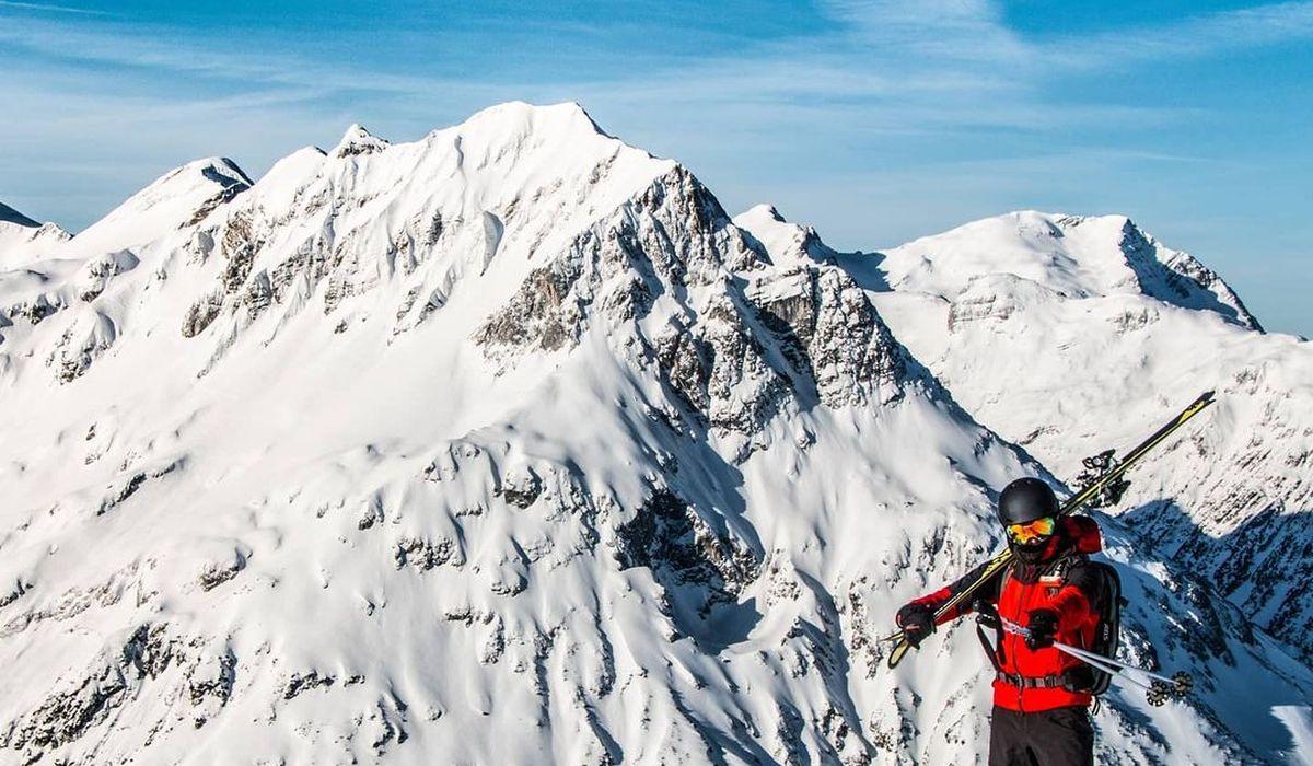 Skier in red jacket stands on snowy mountain peak with skis, surrounded by majestic snow-covered mountains under a clear blue sky.