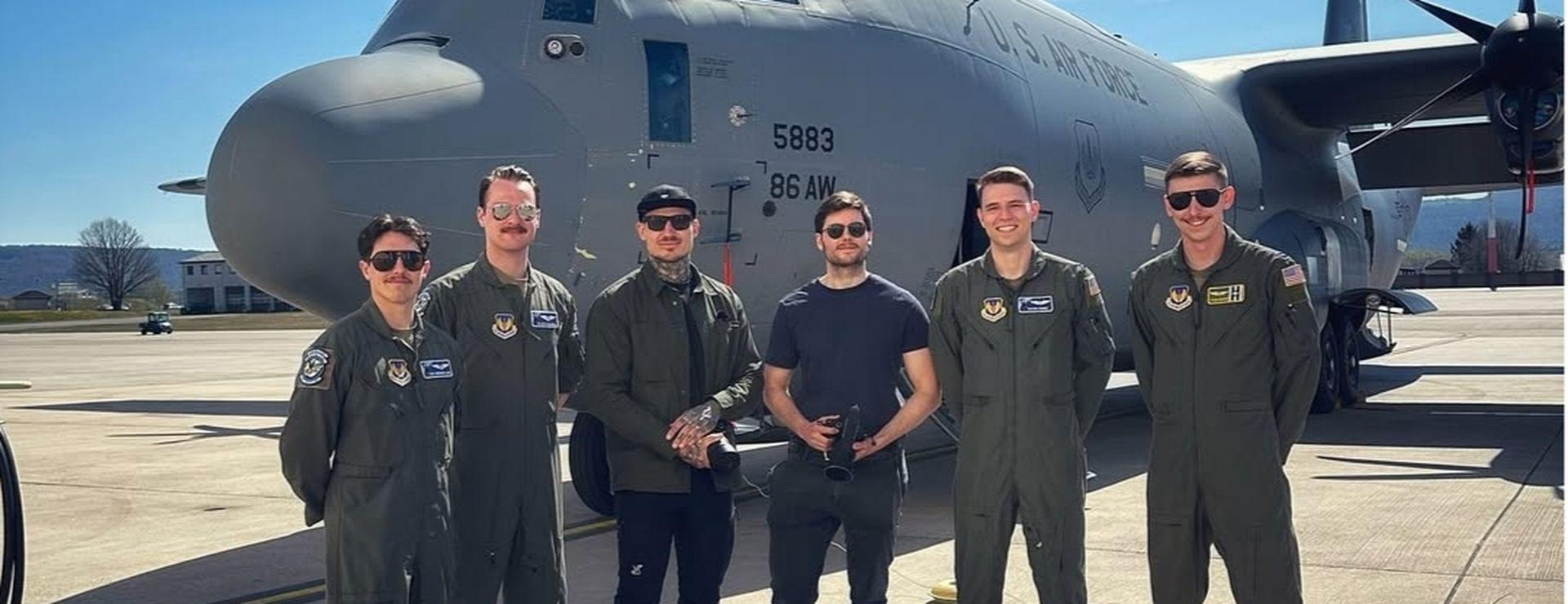 Five people in flight suits stand in front of a large military aircraft on a sunny day at an airbase.