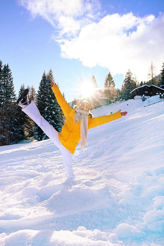 Person in a yellow coat and white pants joyfully poses on a snowy hill with trees and a bright sun in the background.