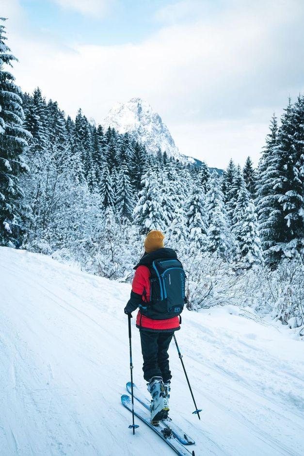 A person skiing uphill on a snowy trail surrounded by snow-covered trees, with a mountain peak visible in the background.