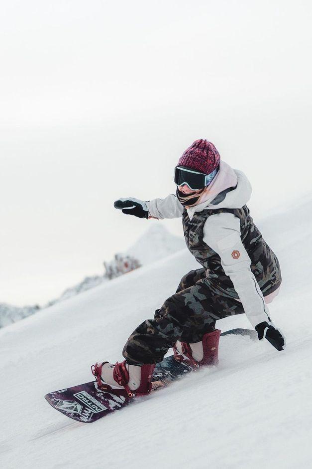 A snowboarder in a pink beanie and camouflage outfit glides down a snowy slope, with mountains in the background.