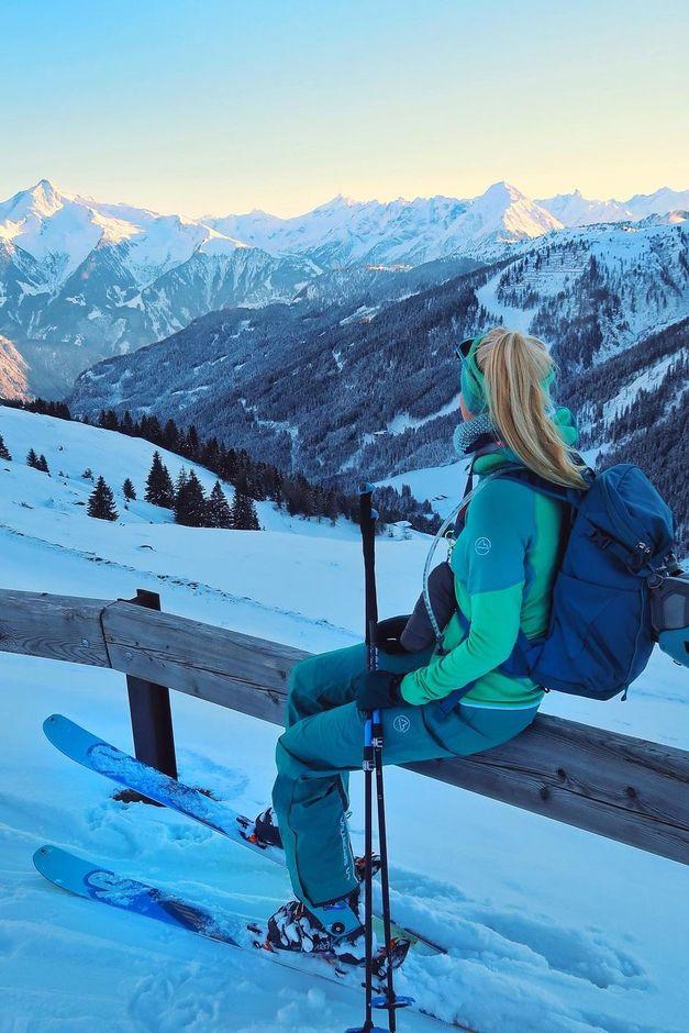 A skier in teal gear sits on a wooden fence, overlooking snow-covered mountains under a clear sky.