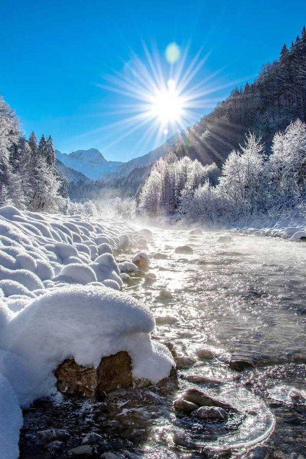 A sunlit winter scene with a river flowing through a snow-covered landscape, surrounded by frosty trees and mountains under a clear blue sky.