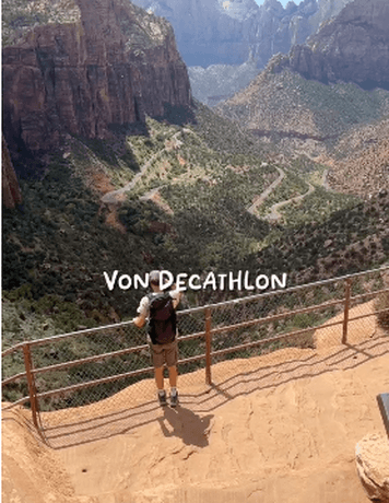 Person standing on a cliff, overlooking a vast canyon landscape with a winding road below under a clear blue sky.