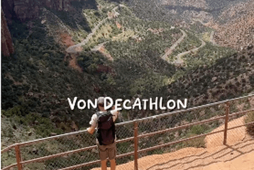 Person standing on a cliff, overlooking a vast canyon landscape with a winding road below under a clear blue sky.