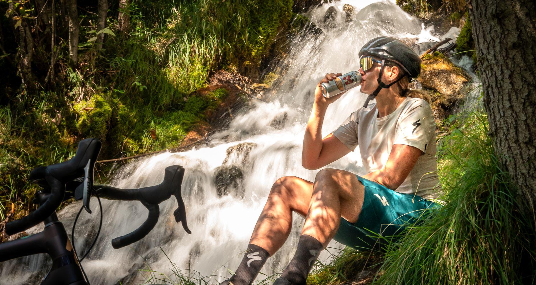 woman sitting next to a waterfall drinking peach flavoured beer