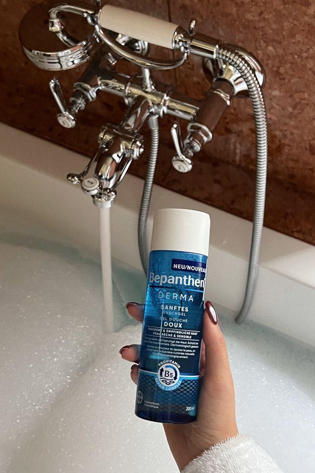 running bathtub in the background and a hand holding a bepanthen derma product in the front