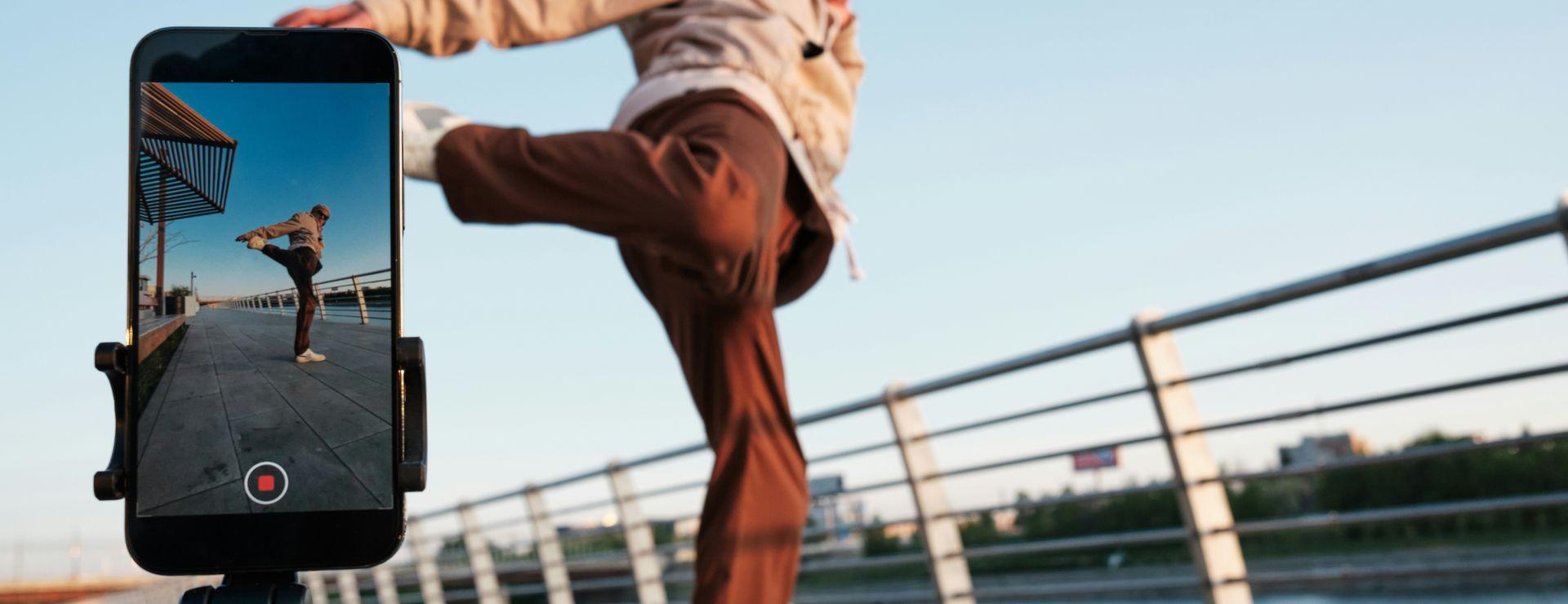 Person in motion, kicking playfully on a riverside walkway, with a phone on a tripod capturing the scene against a blue sky.