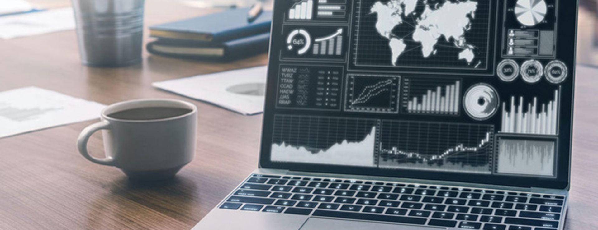 Laptop on a desk displaying data visualizations and world map. Nearby are a coffee cup, notebook, and pen.