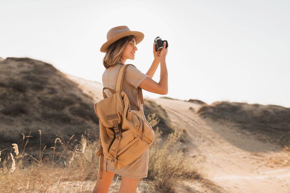 A woman in a hat and dress stands in a desert landscape, holding a camera, with a backpack on her back and hills in the background.
