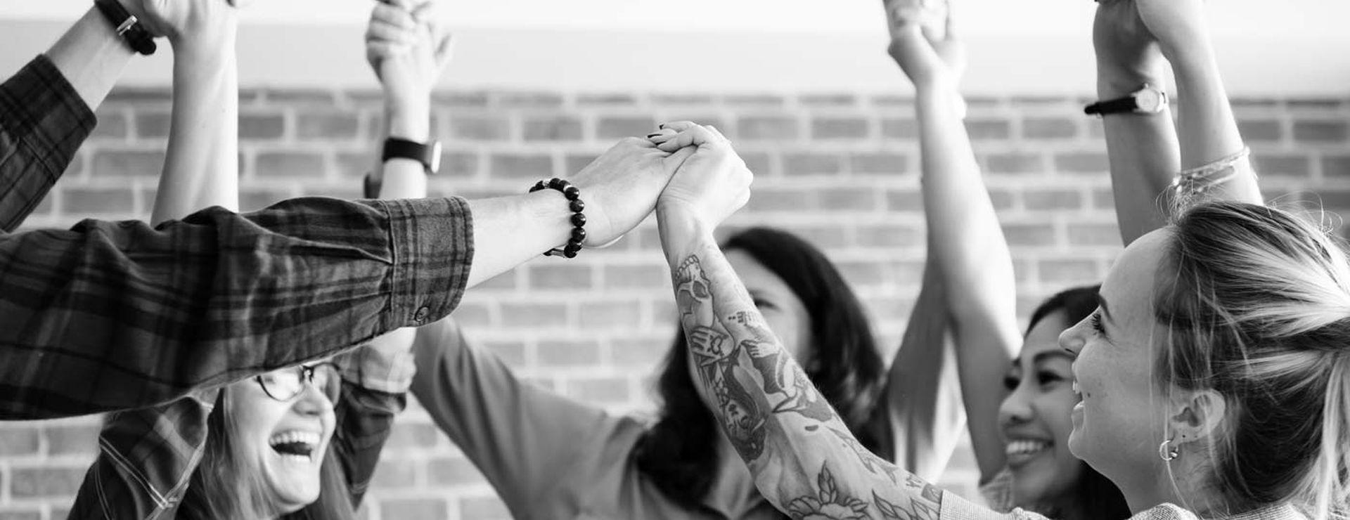 A group of people joyfully raising their hands together in a circle, set against a brick wall background.