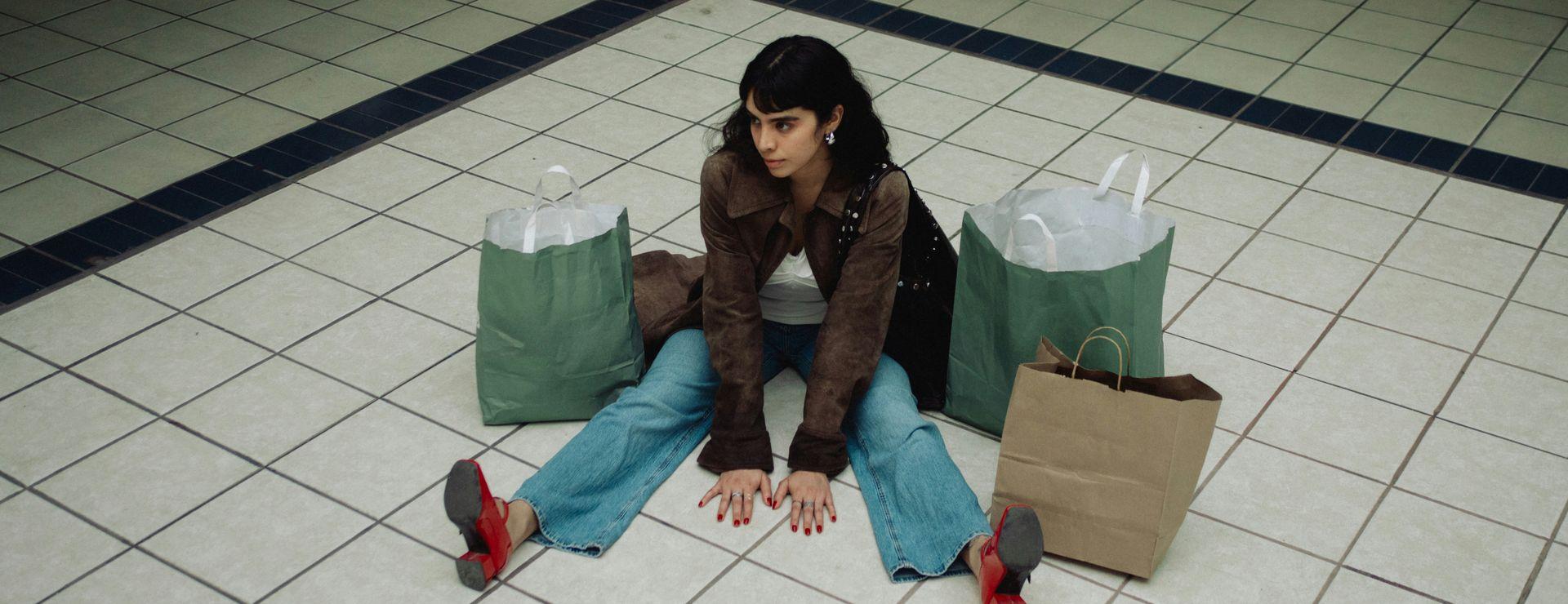 Person sitting on a tiled floor with legs outstretched, surrounded by three shopping bags, wearing red shoes, blue jeans, and a brown jacket.